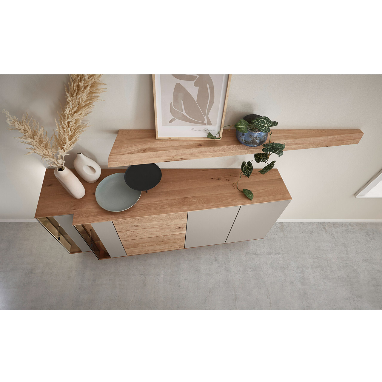 Hartmann Möbel SVEA - Sideboard 8660-4202 / Wandboard 1188 - Kerneiche Natur, Lack Sand, Riffholz - Milieubild Von oben dargestellte Kommode aus massiver Kerneiche mit Lackfronten Sand und Riffholz. Mit zwei Lacktüren, einem Schubkasten und Tür Riffholz und zwei Glas-Lacktüren in asymmetrischem Design und beleuchteter Riffholzrückwand. Darüber Riffholz-Wandboard.