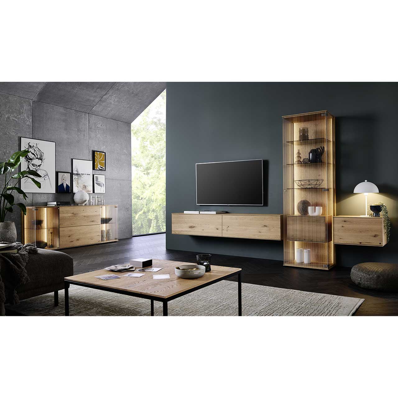 Wohnzimmer mit dreiteiliger Wohnwand aus Kerneiche Natur: zwei Hängelowboards und eine Standvitrine vor dunkelgrauer Wand. Links beleuchtetes Sideboard mit Glastüren, im Vordergrund rechteckiger Couchtisch.