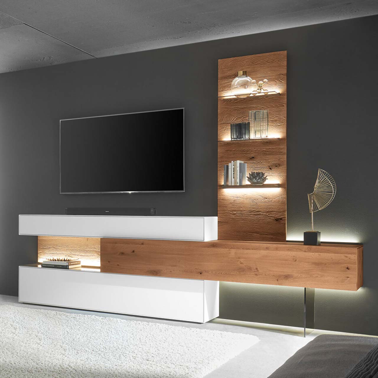 Moderne Wohnwand aus weiß lackierter Birke und dunkler Kerneiche Umato bestehend aus einem Hängelowboard seitlich eingefasst in einen weißen Ober - und Unterschrank mit Beleuchtung und einem Wandpaneel mit drei beleuchteten Glasablageböden. 