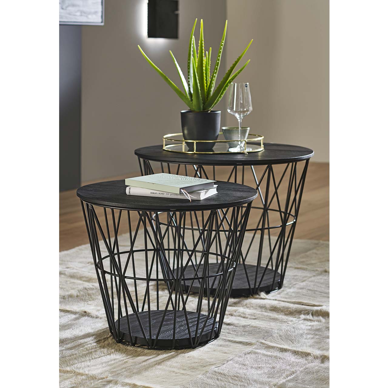 Zwei raffinierte, runde Beistelltische mit feinem Metallkorb in Anthrazit und abnehmbarem Holzdeckel sowie Bodenplatte aus Kernesche anthrazit – im Wohnzimmer stehend.