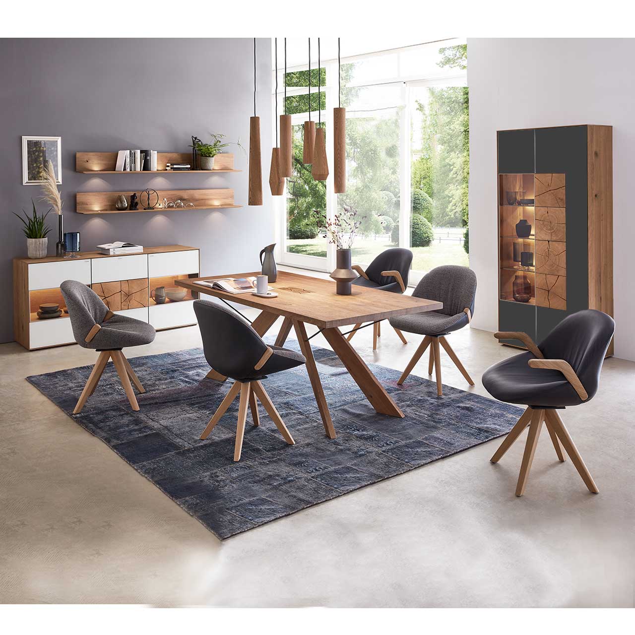 Milieubild Esszimmer - Hartmann CAYA - Standvitrine 7170-0093A re. Esszimmer mit massivem Holztisch und fünf Stühlen mit Holzgestell, rechts an der Wand stehend eine Strandvitrine in Mattglas anthrazit und beleuchtetem Glasausschnitt mit drei Glasböden. Links das passende Sideboard und zwei Wandregale.