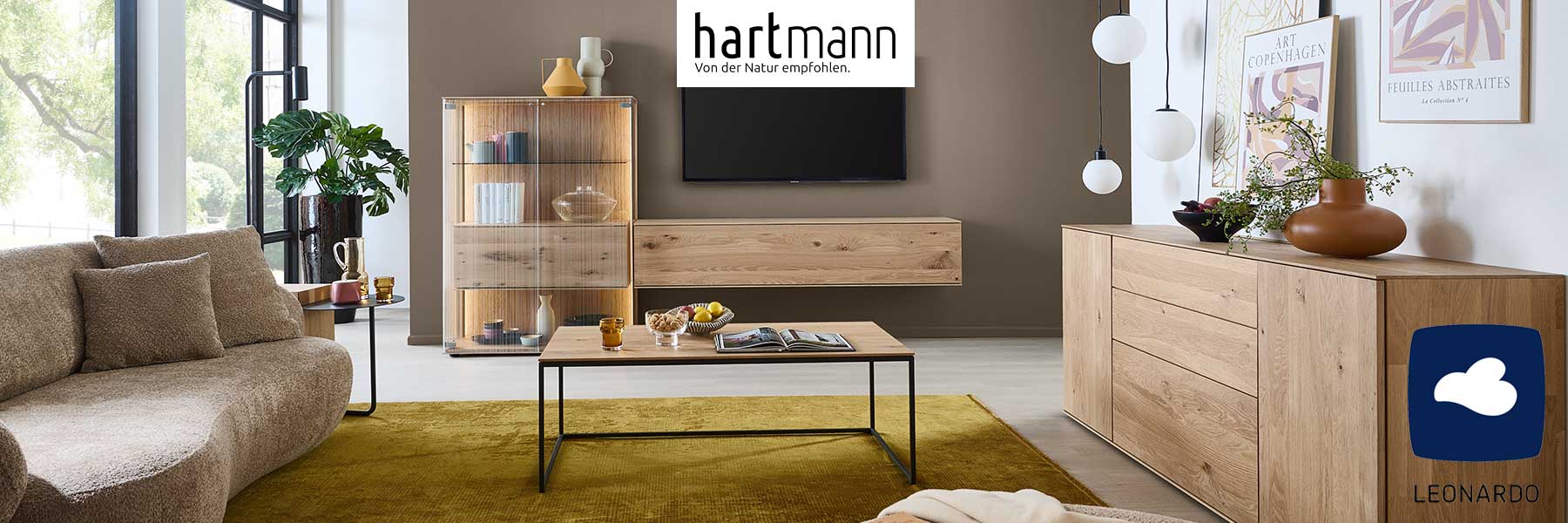 Hartmann Möbel - Möbelserie LINEA Leonardo Living - Kerneiche Natur Hartmann LINEA - Massivholzmöbel aus Kerneiche Natur kombiniert mit satiniertem Glas. Ambientebild zeigt schlichtes Hängelowboard, links eine Standvitrine edlen Glasfronten, davor minimalistischer Couchtisch, rechts langes, schlichtes Sideboard.