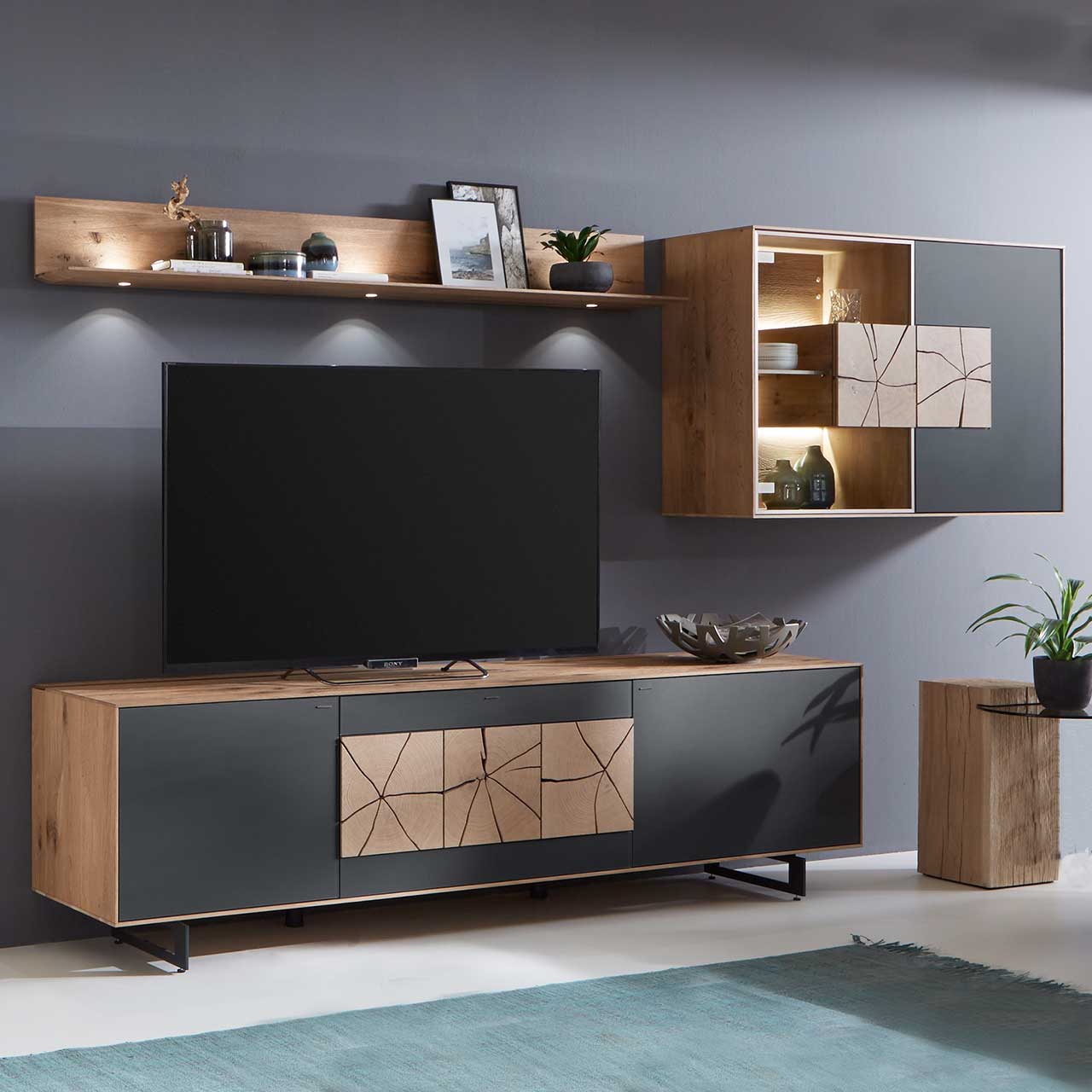Moderne dreiteilige Massivholz Wohnwand aus Kerneiche Umato mit Mattglas anthrazit Fronten: Lowboard mit dekorativem Hirnholz auf Metallkufen, Wandregal aus Holz, Hängevitrine mit beleuchtetem Glasausschnitt und Hirnholz.