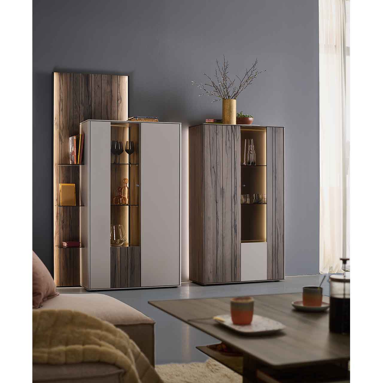Milieubild - Hartmann AUDRA - Highboards 5610-8092 und 8095 - Altholzbuche Uyaa & Lack Sand Wohnzimmer mit zwei modernen Massivholz-Highboards in sandfarbenen Lack und dunkelbrauner Altholzbuche Uyaa, jeweils mit beleuchtetem Glasausschnitt und Glasböden, ergänzt durch ein seitliches, beleuchtetes Holzwandpaneel.