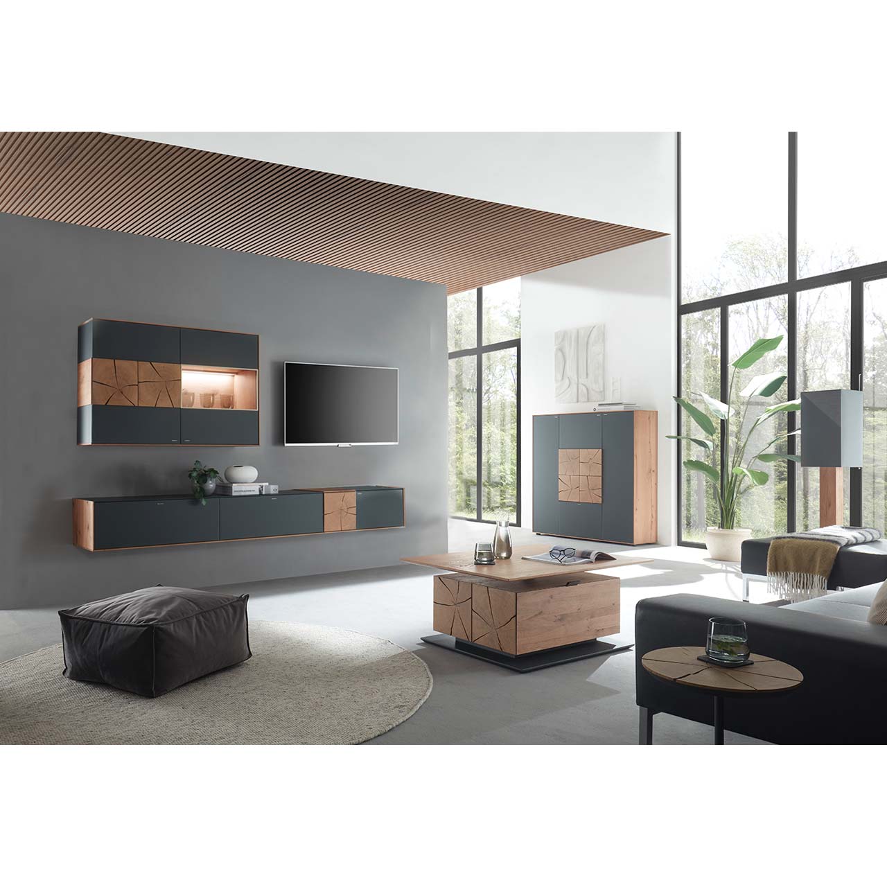 Wohnzimmer mit TV und moderner Wohnwand aus massiver Kerneiche und Mattglas anthrazit Fronten, mit schmalem Hängelowboard und beleuchteter Hängevitrine. Rechts das passende Highboard. Im Vordergrund ein Holz-Couchtisch mit rechteckiger Glasplatte.