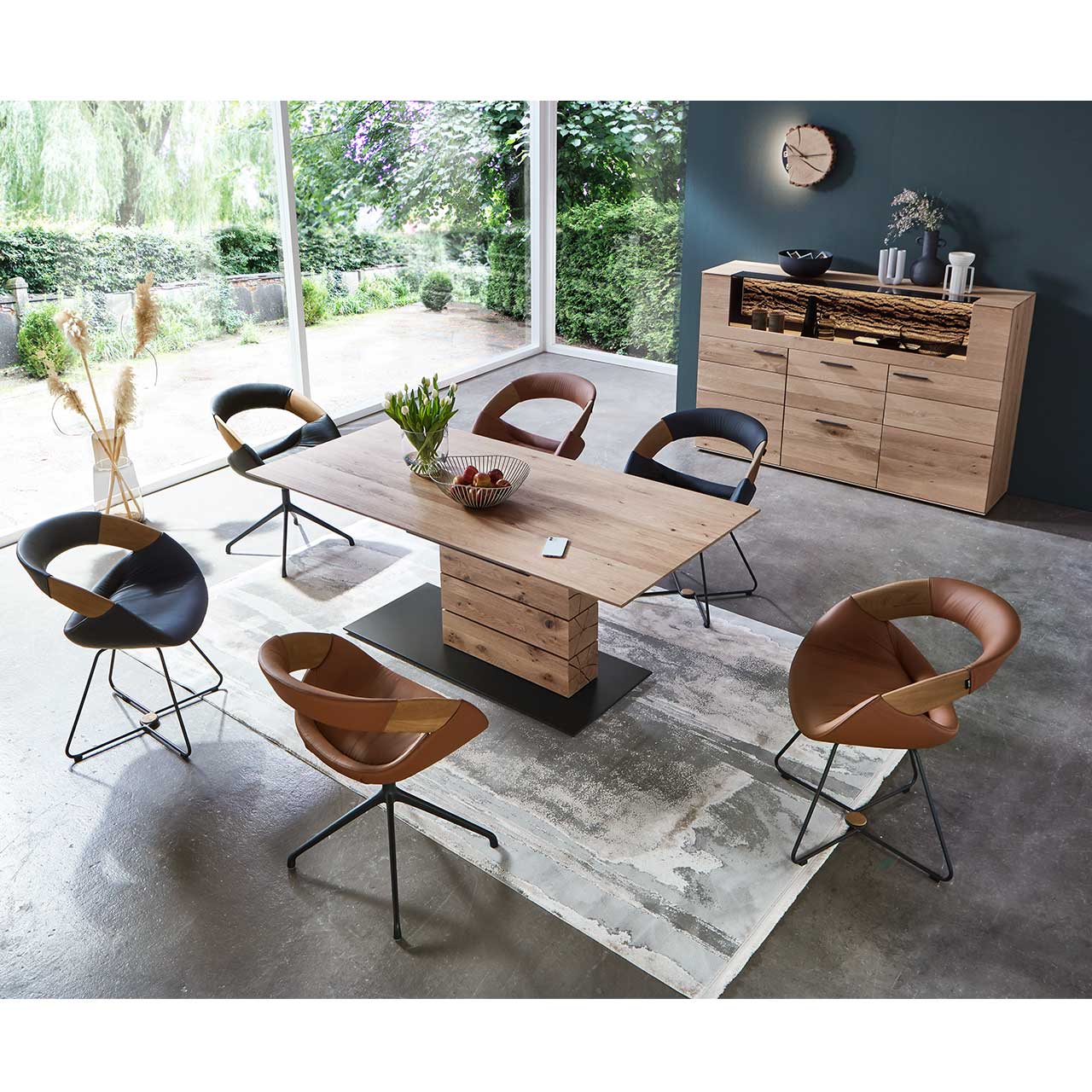 Blick von oben auf ein modernes Esszimmer mit massivem, rechteckigem Holztisch, umgeben von sechs Stühlen auf Drahtgestell. Rechts vor dunkler Wand das passende Highboard aus Kerneiche mit Glasfront.