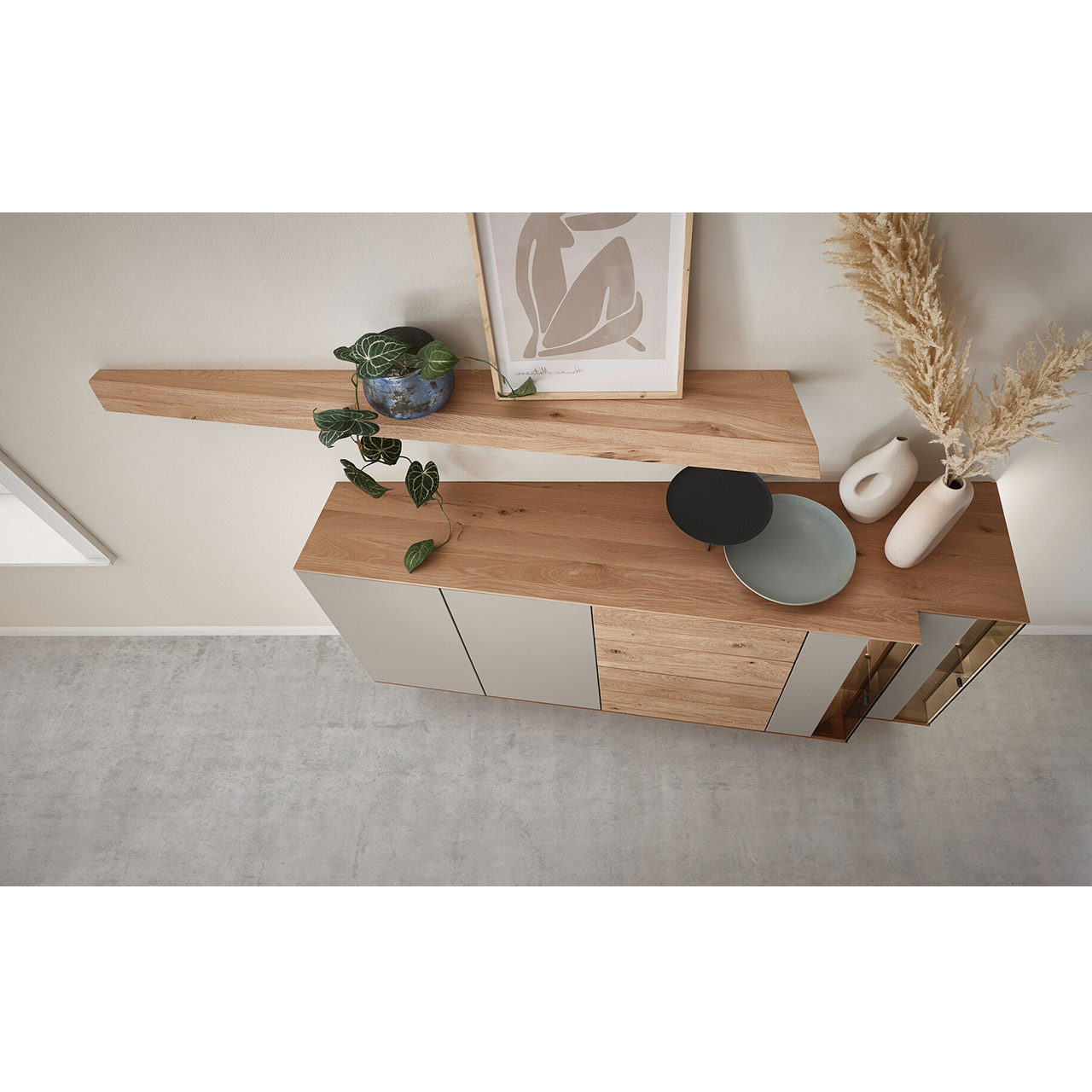 Von oben dargestellte Kommode aus massiver Kerneiche mit Lackfronten Sand und Riffholz. Mit zwei Lacktüren, einem Schubkasten und Tür Riffholz und zwei Glas-Lacktüren in asymmetrischem Design und beleuchteter Riffholzrückwand. Darüber Riffholz-Wandboard.