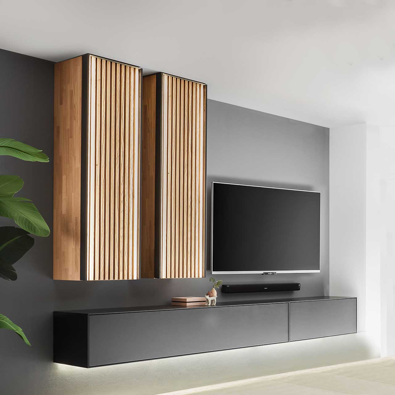 Hartmann SELINO - Wohnwand 8560 Nr. V20 Wohnzimmer mit Fernseher und moderner vierteiliger Wohnwand aus massivem Holz: Zwei schlichte Hängelowboards anthrazit lackiert und zwei hohe Hängeschränke mit hellen Holzlamellen – vor dunkelgrauer Wand.