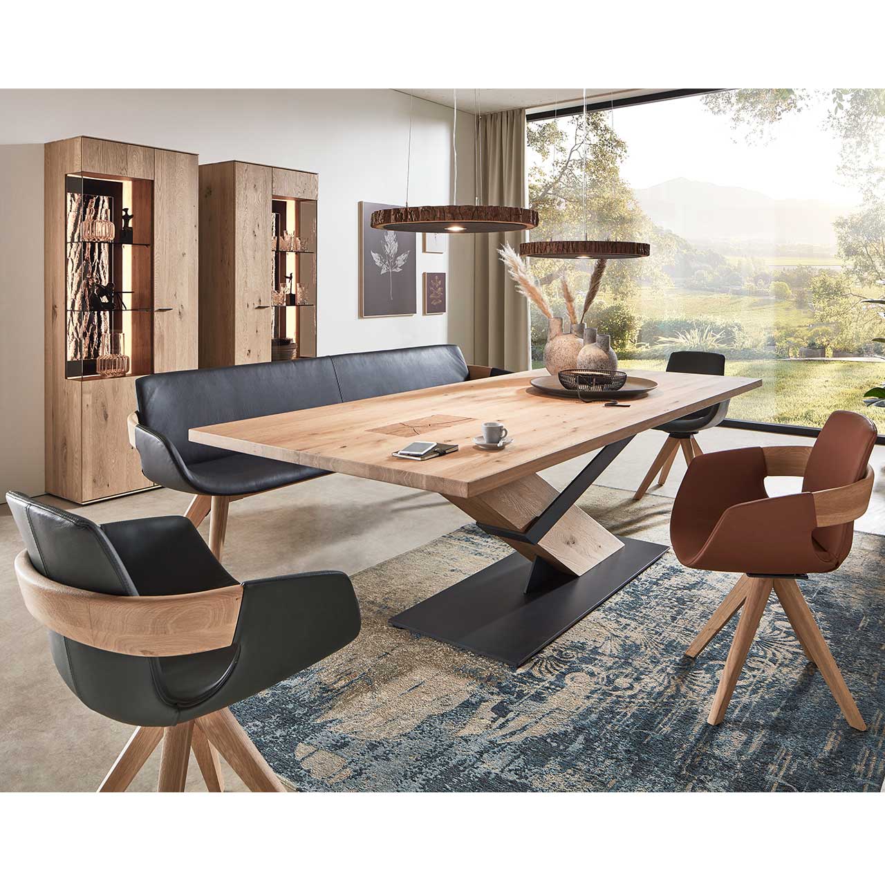 Modernes Esszimmer mit massivem Holztisch, drei Drehstühlen auf Holzgestell und einer Sitzbank in schwarzem Leder. Über dem Tisch hängen zwei Tellerleuchten aus echter Holzscheibe. Dahinter an der Wand zwei passende Standvitrinen.