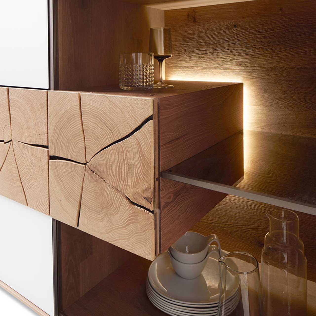 Hartmann CAYA - Hängeelement 7170-4135W li. Hartmann Detailbild eines beleuchteten Hirnholzblocks aus massiver Kerneiche Umato mit sichtbarer Holzmaserung und charakteristischen Holzrissen an Glastür eines CAYA Sideboards oder einer Hängevitrine. Möbel Caya - Sideboard 7170-4195 W - Detail