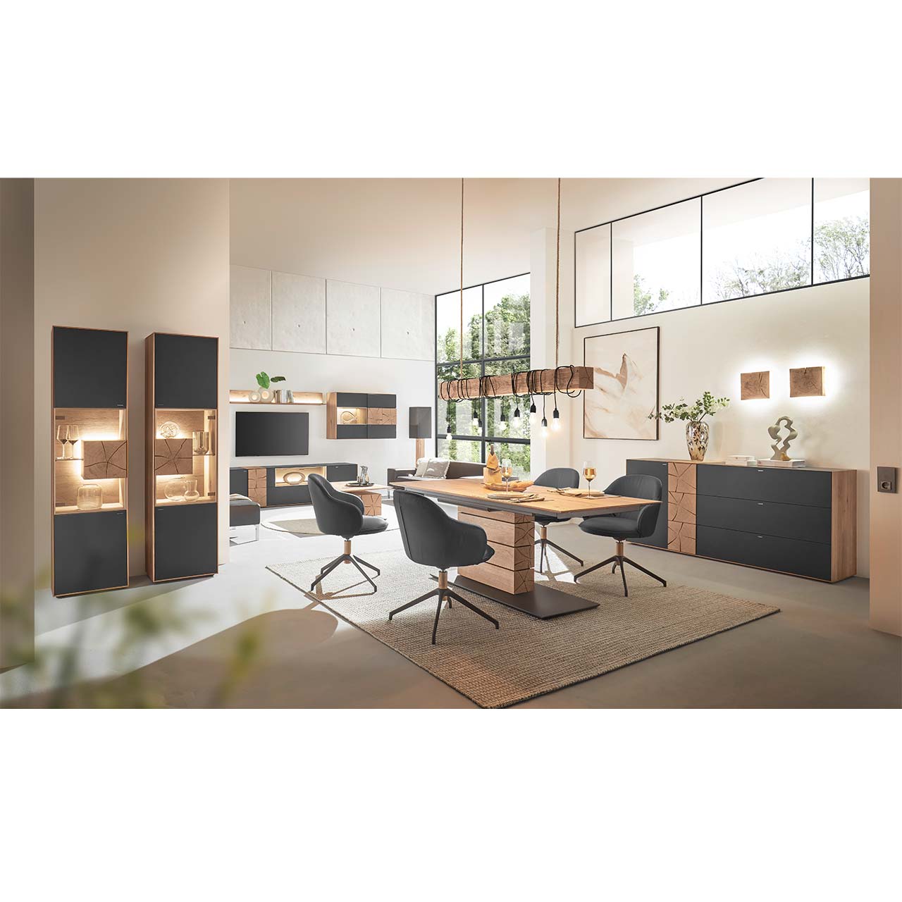 Modern eingerichtetes Esszimmer mit Esstisch aus massivem Holz umgeben von vier Stühlen mit schwarzem Lederbezug und Metallgestell. Darüber passende Balkenleuchte. Links ein Sideboard mit Mattglas Anthrazit Front, rechts zwei passende Standvitrinen.