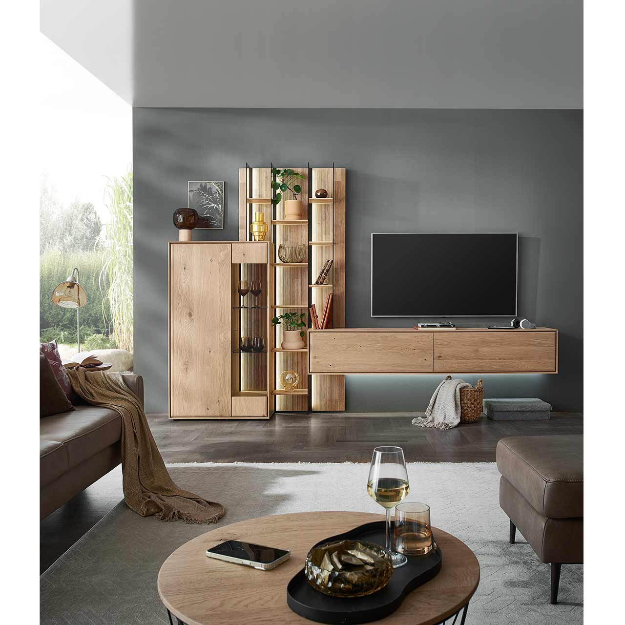Modernes Wohnzimmer mit dreiteiliger Massivholzwohnwand bestehend aus Highboard, hohem Wandregal mit Holzböden, beleuchteten Glasseiten und geschroppter Rückwand und einem darin integriertem Hängelowboard. Davor runder Couchtisch mit Sessel.