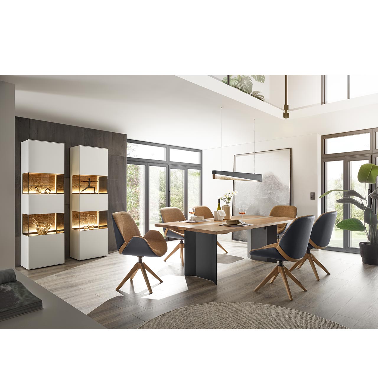 Modernes Esszimmer mit rechteckigem Esstisch aus hellem Holz mit 2-Fuß-Holzuntergestell in Anthrazit. Umgeben von sechs modernen Stühlen in beigem Stoff, mit Außensitzschale und Metallgestell in anthrazit. Links zwei Standvitrine in Lack weiß.