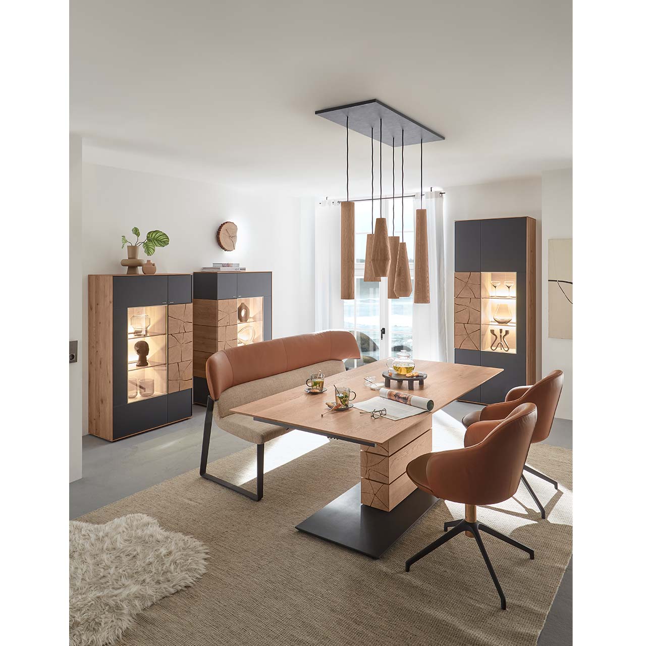 Modernes Esszimmer mit rechteckigem Massivholz Tisch mit Sitzbank und zwei Stühlen mit Lederbezug in cognac. Über dem Tisch hängt ein Pendelleuchte mit Holz-Lampenschirmen. Im Hintergrund stehen zwei passende Highboards und eine Standvitrine in Anthrazit.