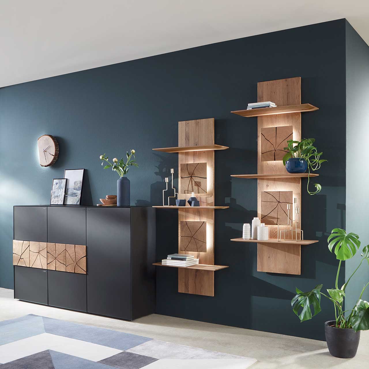 Zwei hohe Regalelemente aus massiver Kerneiche Umato, mit je drei Ablageböden und je zwei beleuchteten, rechteckigen Hirnholzscheiben. Links daneben ein modernes Sideboard mit Mattglas Anthrazit Front und vier Hirnholzzscheiben - vor dunkler Wand.