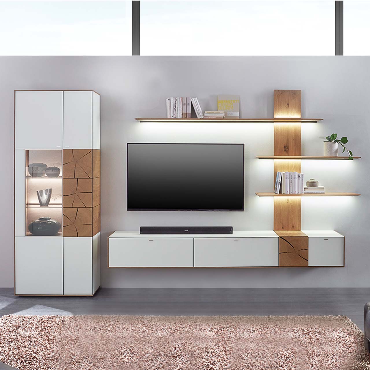 Hartmann CAYA - Wohnwand 7170 Nr. V74W - dreiteilig Massivholz Wohnwand aus Kerneiche mit Mattglas weißen Fronten. Unten ein Hängelowboard mit dekorativer Holzapplikation, seitlich ein Wandpaneel mit Regalböden und indirekter Beleuchtung, links passende Standvitrine, dazwischen hängender Fernseher.