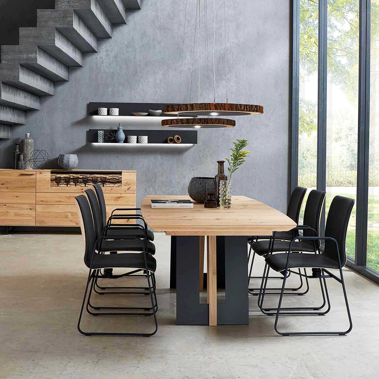 Modernes Esszimmer mit massivem, rechteckigem Holztisch und sechs passenden Stühlen mit schwarzem Leder und Metallgestell. Darüber hängend zwei Baumscheiben-Leuchten. Im Hintergrund die passenden Esszimmermöbel.