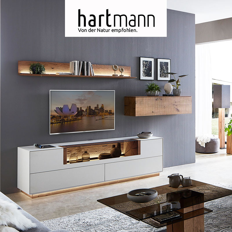 Hartmann BENJO - Wohnzimmermöbel aus massiver Birke weiß lackiert kombiniert mit Kerneiche Umato. Ambientebild zeigt beleuchtetes Holzpaneel, weißes Lowboard mit Glasklappe und Sockel und einen Hängeschrank aus massiver Kerneiche - vor grauer Wand.  