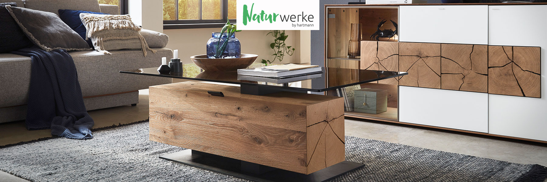 Hartmann NATURWERKE - individuelle und funktionelle Couchtische aus Massivholz kombiniert mit Glas und Metall. Ambientebild zeigt Couchtisch mit edler Glasplatte in Bronze und eckiger Holzsäule als Stauraumfach aus massiver Kerneiche Natur.