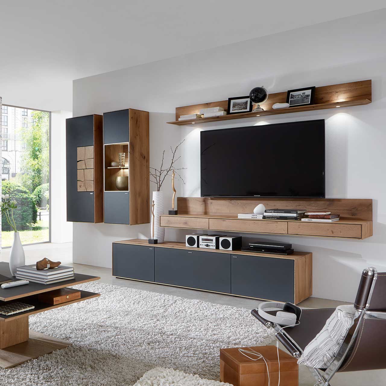 Wohnzimmer mit TV und moderner Wohnwand aus massiver Kerneiche Umato mit Mattglas anthrazit Fronten: Beleuchtetes Wandregal, langes Lowboard, beleuchtete Hängevitrine und Hängeschrank mit dekorativen Hirnholzscheiben.