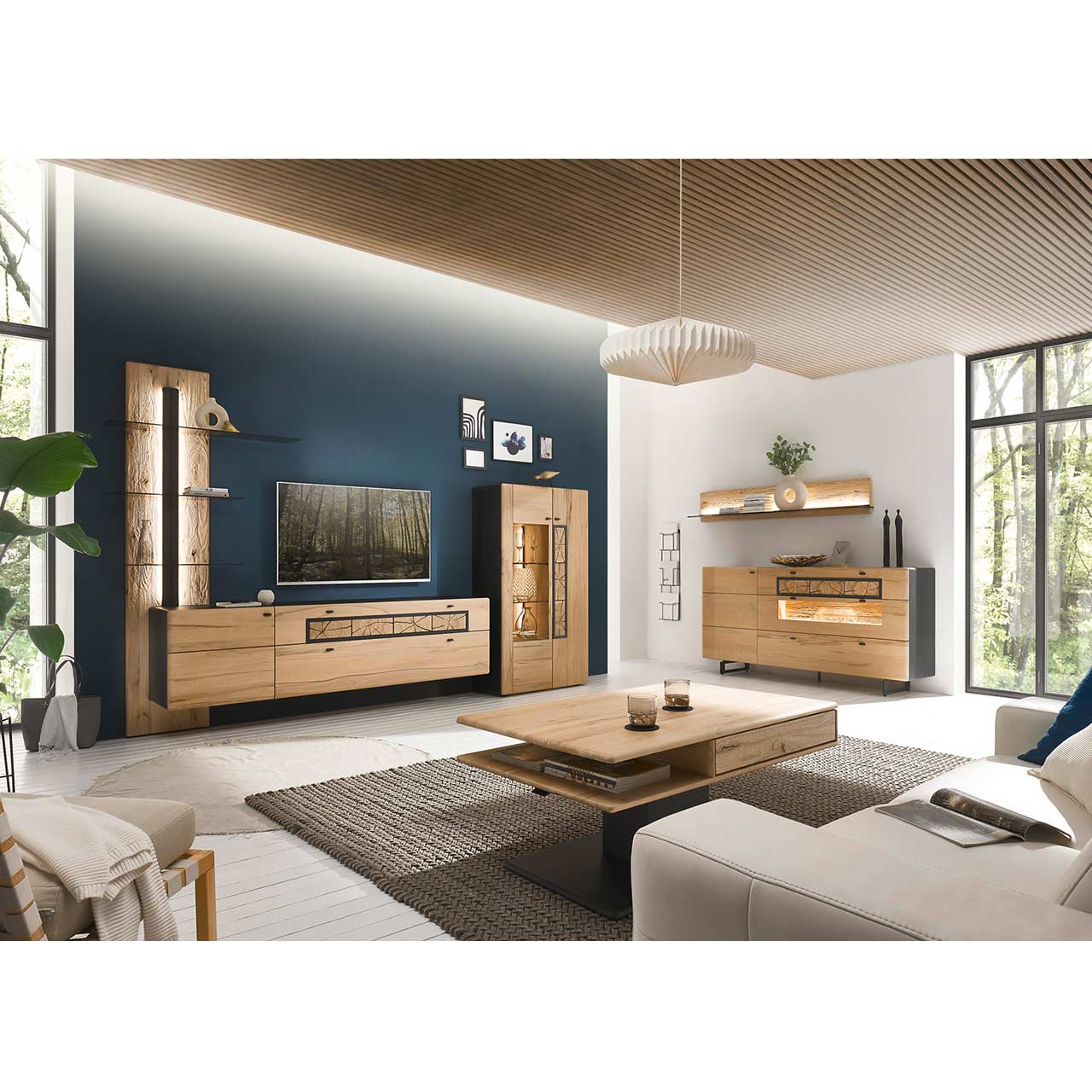 Eingerichtetes Wohnzimmer mit moderner Wohnwand aus Kernbuche vor einer dunkelblauen Wand bestehend aus Highboard, Lowboard und Paneel. Links das passende Sideboard auf Metallkufen darüber ein Regal. Im Vordergrund der dazugehörige Couchtisch.