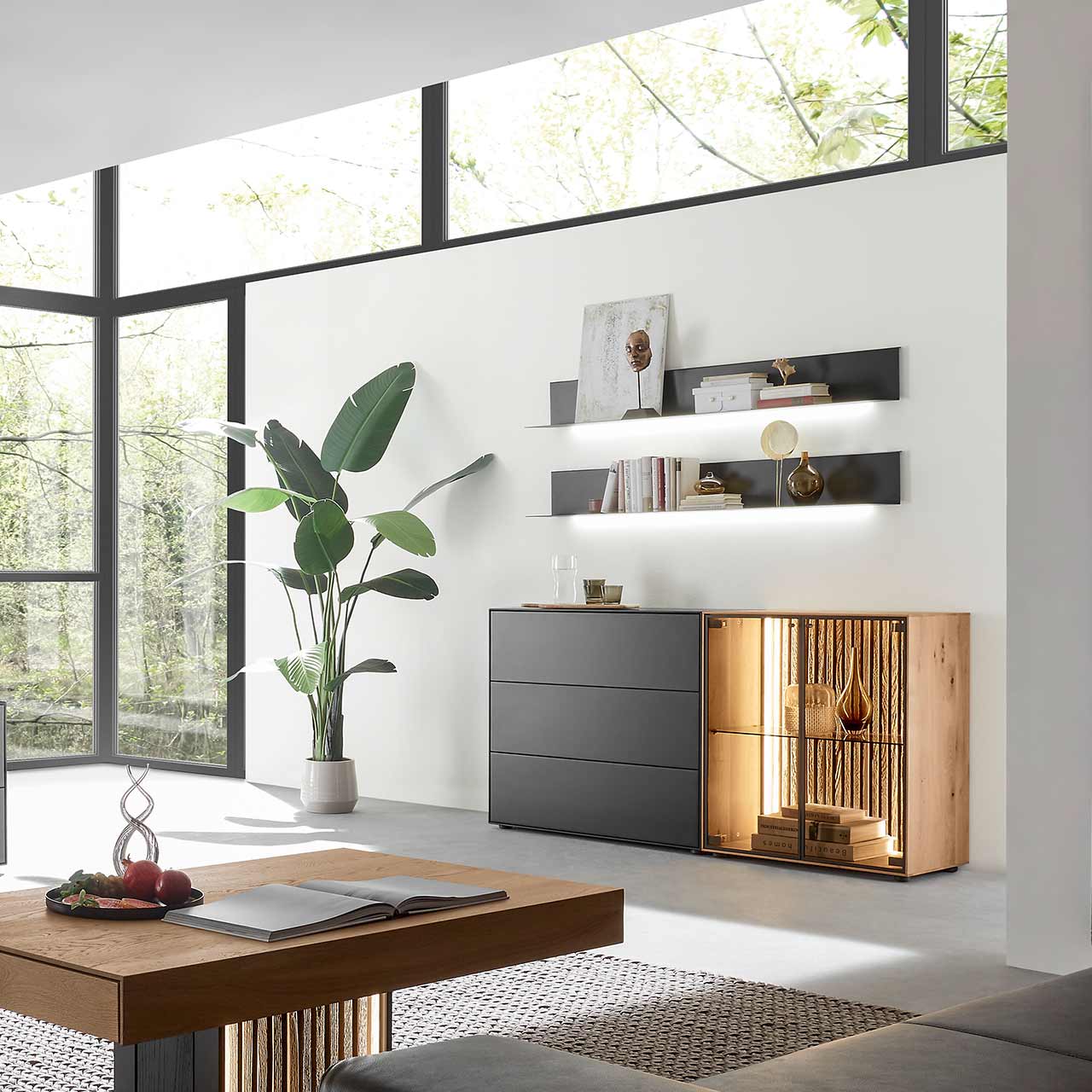 Modernes Wohnzimmer mit beleuchtetem Sideboard aus Birke anthrazit mit Glastüren, Lamellenrückwand und dunklen Schubkästen, darüber hängend zwei Metallregale in anthrazit. Im Vordergrund der passende Holz-Couchtisch.