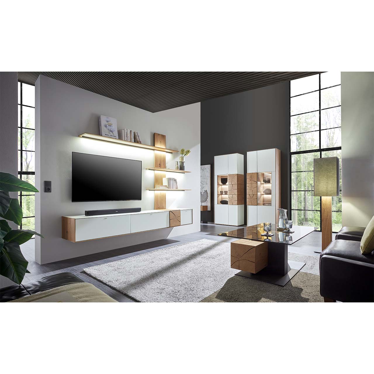 Wohnzimmer mit TV und moderner Wohnwand aus massiver Kerneiche Umato mit  Mattglas weiß Fronten und Hirnholzscheiben, mit Hängelowboard und Wandpaneel mit drei Regalböden. Rechts zwei passende Standvitrinen. Im Vordergrund Couchtisch mit Glasplatte.