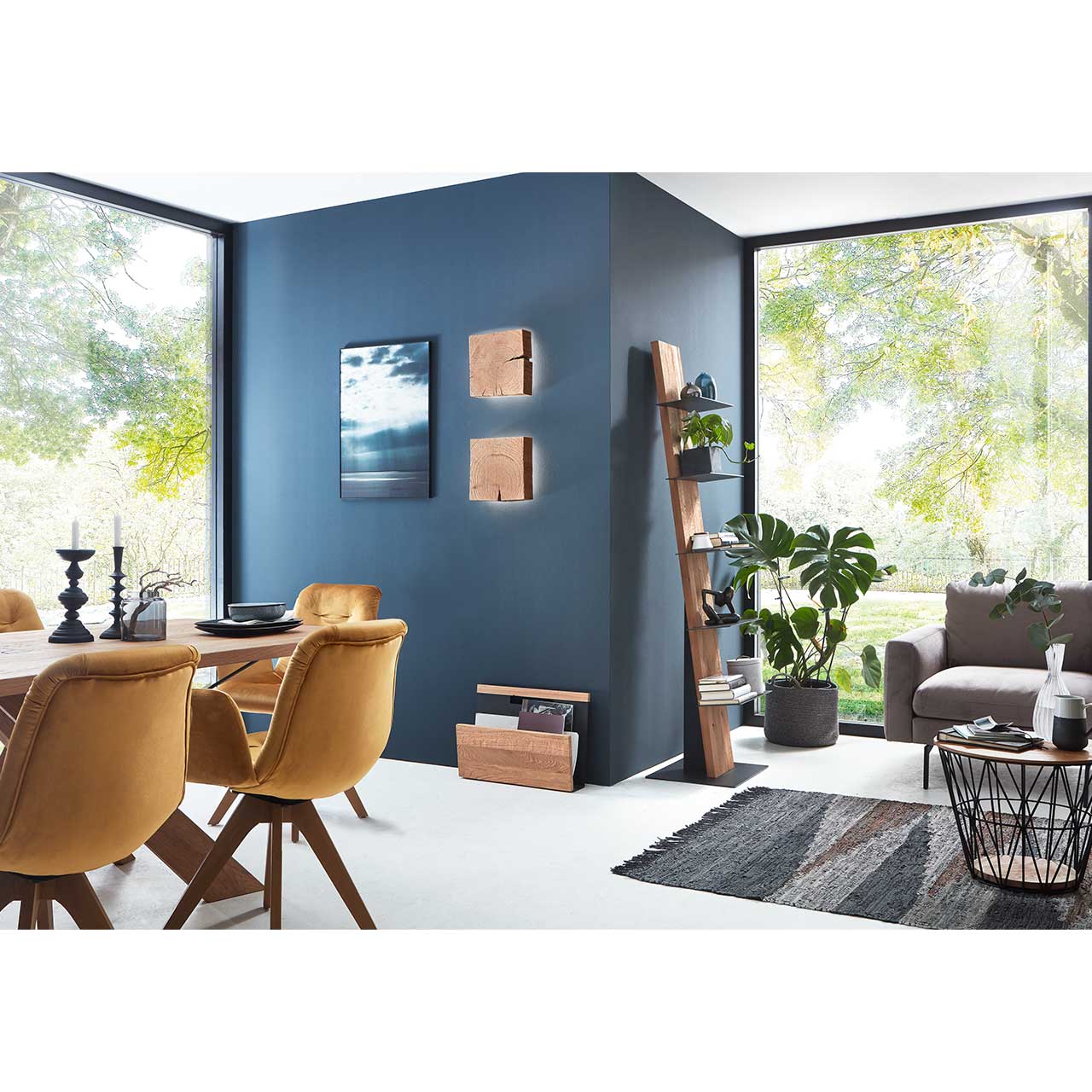Modernes Esszimmer mit Holz-Zeitungsständer, zwei Wandlampen aus Massivholzblock und passendem Stehregal mit Ablagen – vor dunkelblauer Wand. Davor ein runder Beistelltisch mit Metallkorb.