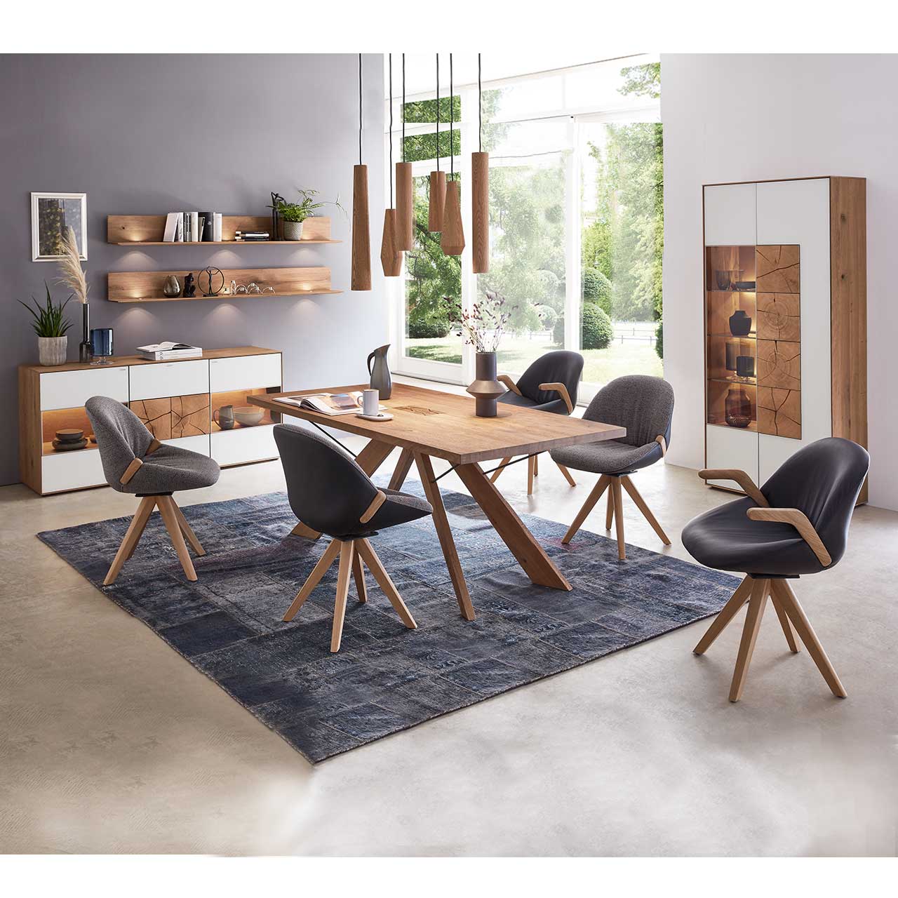 Esszimmer mit massivem Holztisch und fünf Stühlen mit Holzgestell, rechts an der Wand stehend eine Strandvitrine in Mattglas weiß und beleuchtetem Glasausschnitt mit drei Glasböden. Links das passende Sideboard und zwei Wandregale.