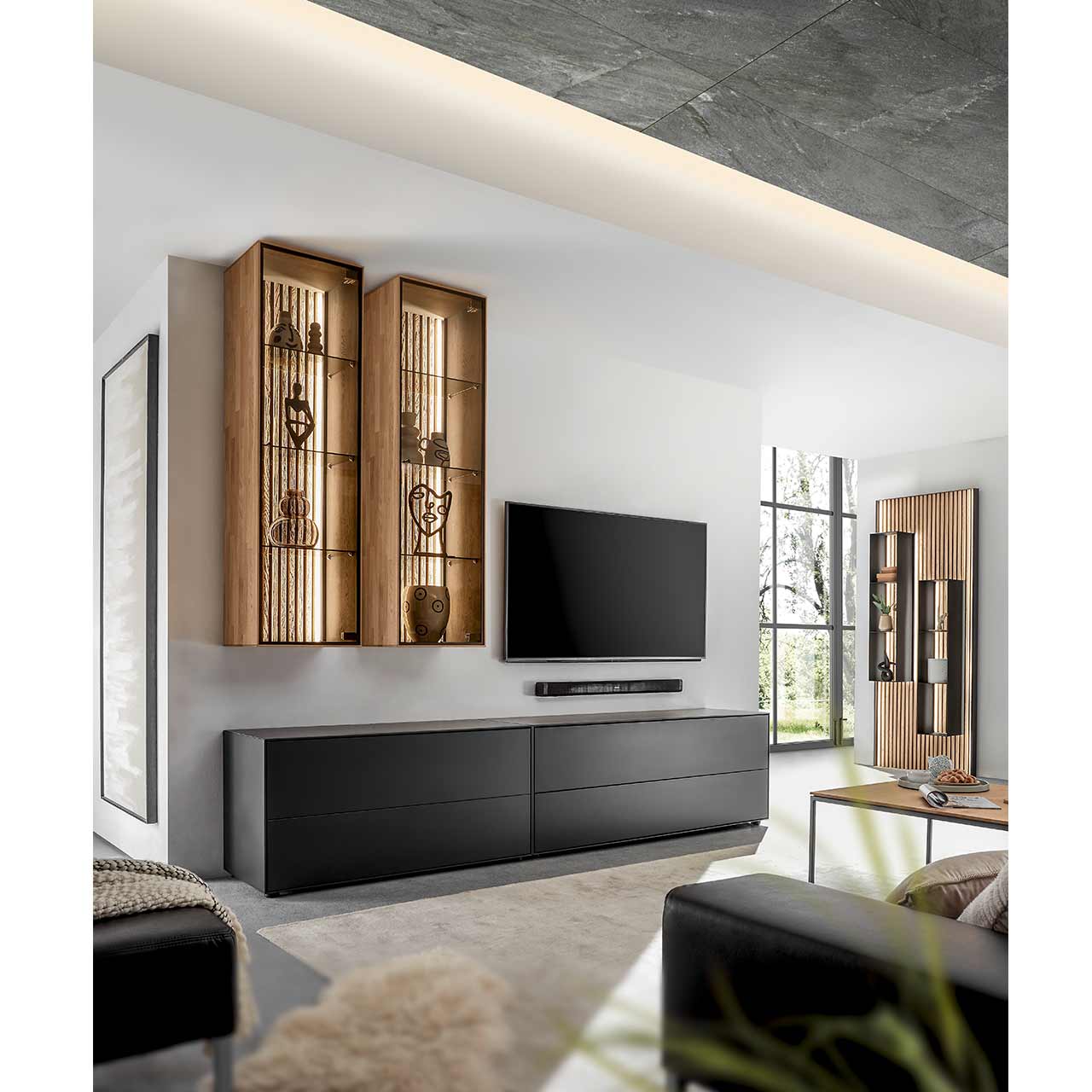 Wohnzimmer mit Fernseher und moderner Massivholz-Wohnwand mit zwei Lowboards aus Birke anthrazit und zwei Hängevitrinen aus heller Kerneiche mit beleuchteter Lamellenrückwand.