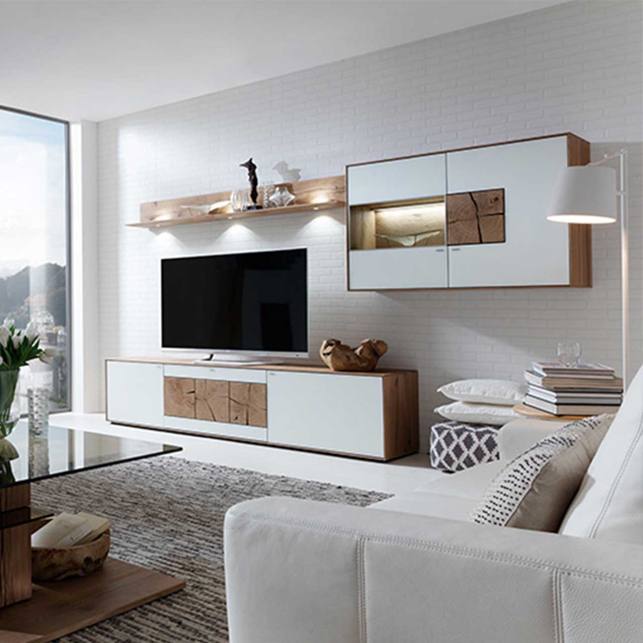 Wohnzimmer mit TV und moderner Wohnwand aus massiver Kerneiche Umato mit Mattglas weiß Fronten: Langes Lowboard mit rechteckigen Hirnholzscheiben, darüber hängend Wandregal aus Holz, daneben beleuchtete Hängevitrine mit Glastür und Holztür.