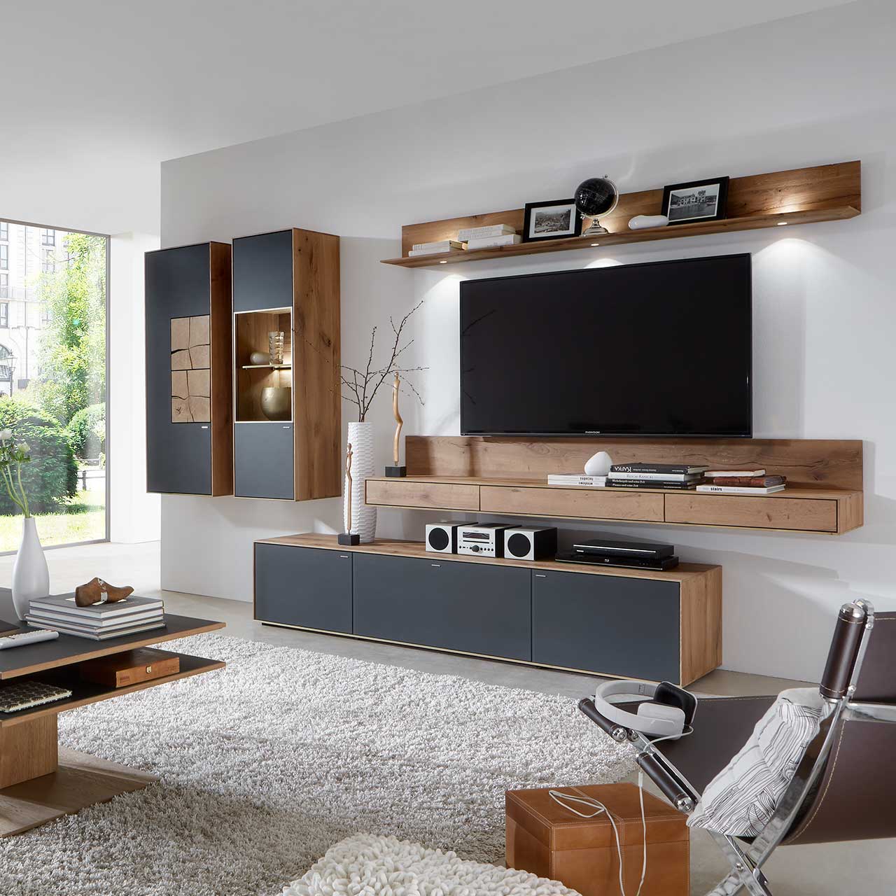 Wohnzimmer mit TV und moderner Wohnwand aus massiver Kerneiche Umato mit Mattglas anthrazit Fronten: Beleuchtetes Wandregal, langes Lowboard, beleuchtete Hängevitrine und Hängeschrank mit dekorativen Hirnholzscheiben.