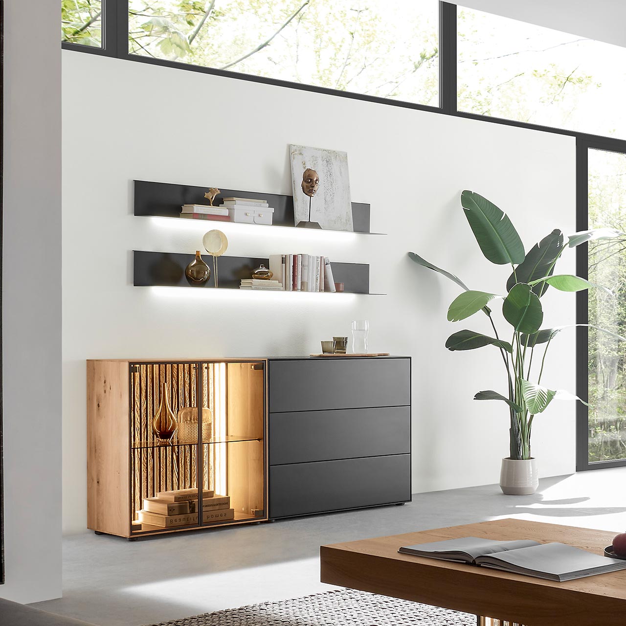Modernes Wohnzimmer mit beleuchtetem Sideboard aus Birke anthrazit mit Glastüren, Lamellenrückwand und dunklen Schubkästen, darüber hängend zwei Metallregale in anthrazit. Im Vordergrund der passende Holz-Couchtisch.