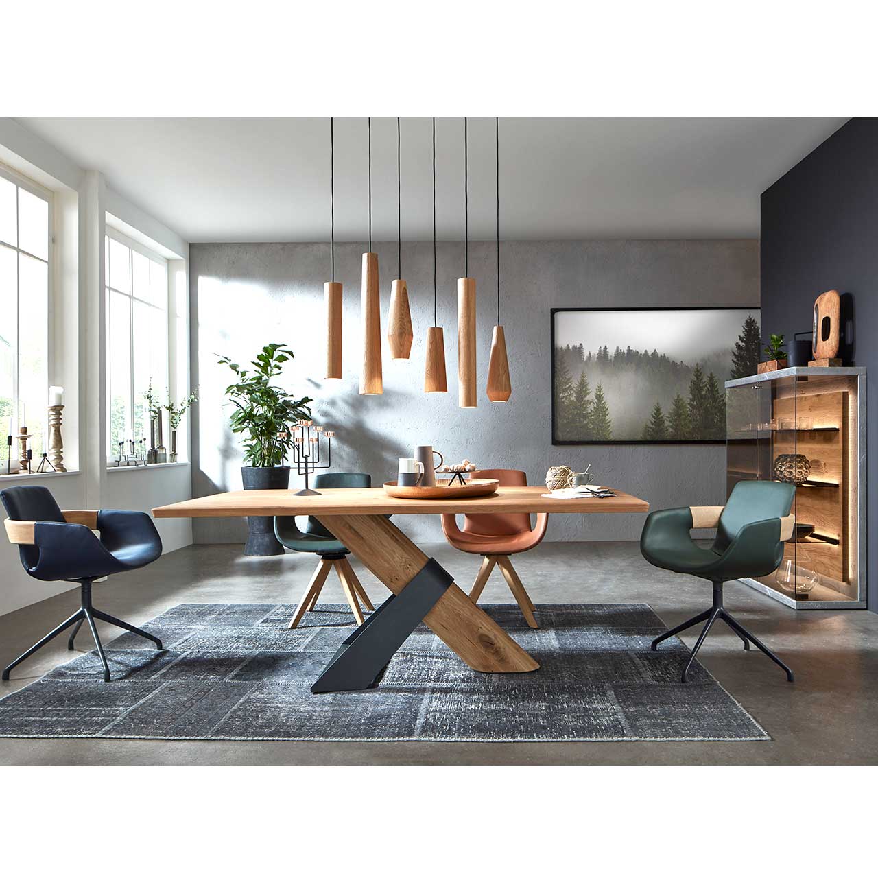 Modernes Esszimmer mit massivem, rechteckigen Holztisch, umgeben von vier Stühlen mit Holz- und Metallgestell. Über dem Tisch hängen sechs Pendelleuchten mit zylinderförmigen Lampenschirmen aus Holz. Links an der Wand ein Glas-Highboard.