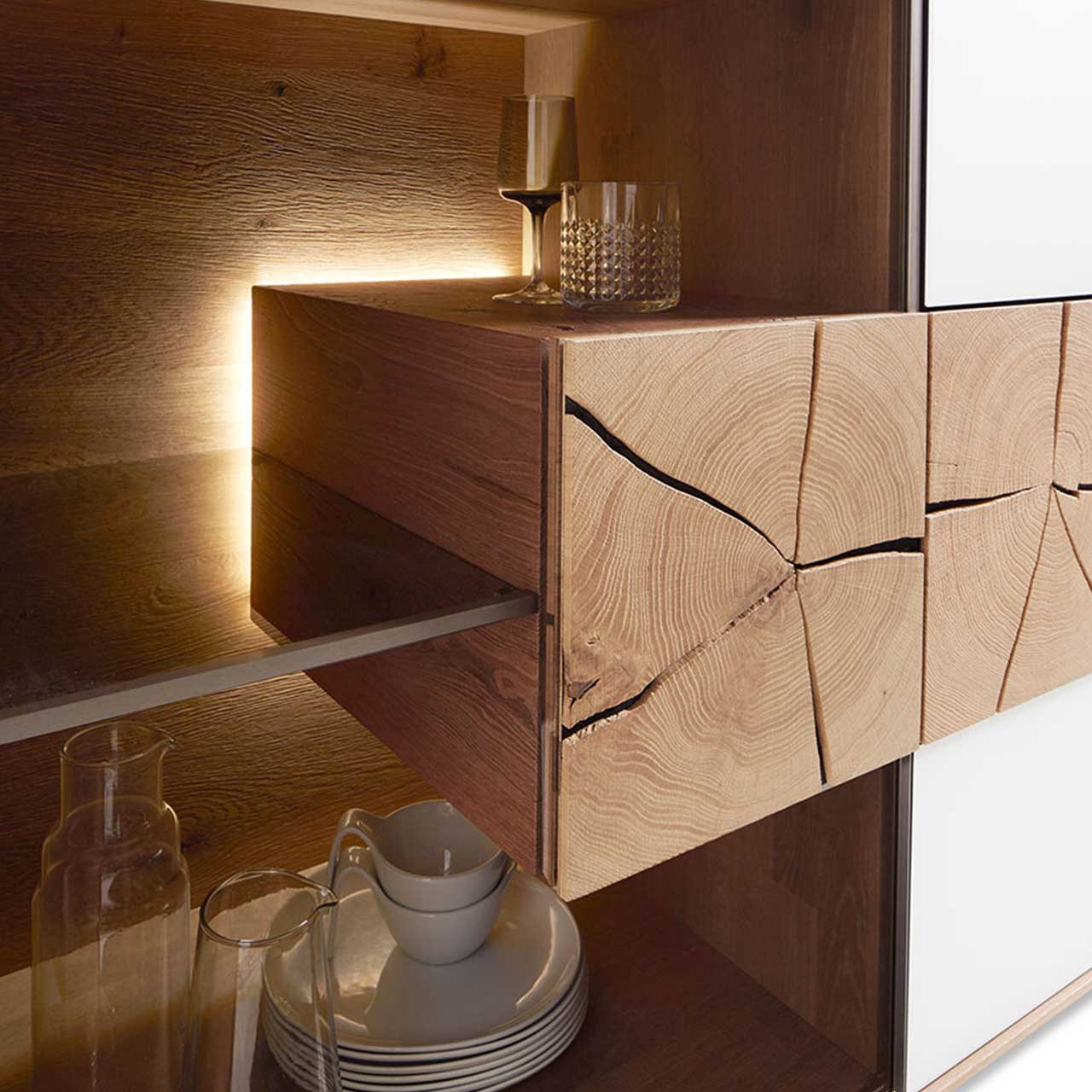 Hartmann CAYA - Hängeelement 7170-4136W re. Detailbild eines beleuchteten Hirnholzblocks aus massiver Kerneiche Umato mit sichtbarer Holzmaserung und charakteristischen Holzrissen an Glastür eines CAYA Sideboards oder einer Hängevitrine.