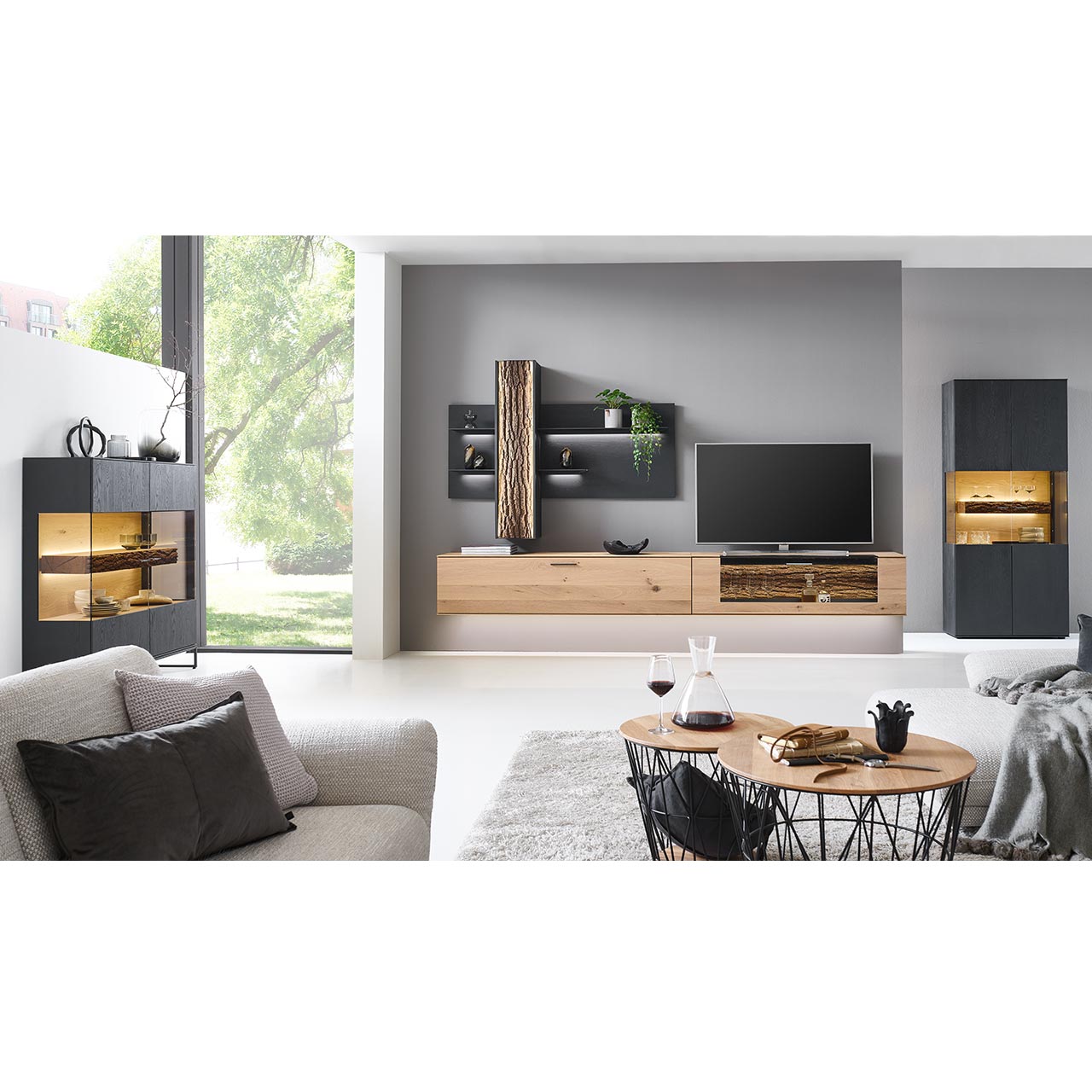 Wohnzimmer mit Wohnwand aus zwei Hängelowboards in massiver Kerneiche Natur, teils verglast, einem Wandpaneel mit beleuchteten Böden und Stauraumschrank, dazu links eine Standvitrine und rechts das passende Highboard in anthrazit gebeizter Kerneiche.