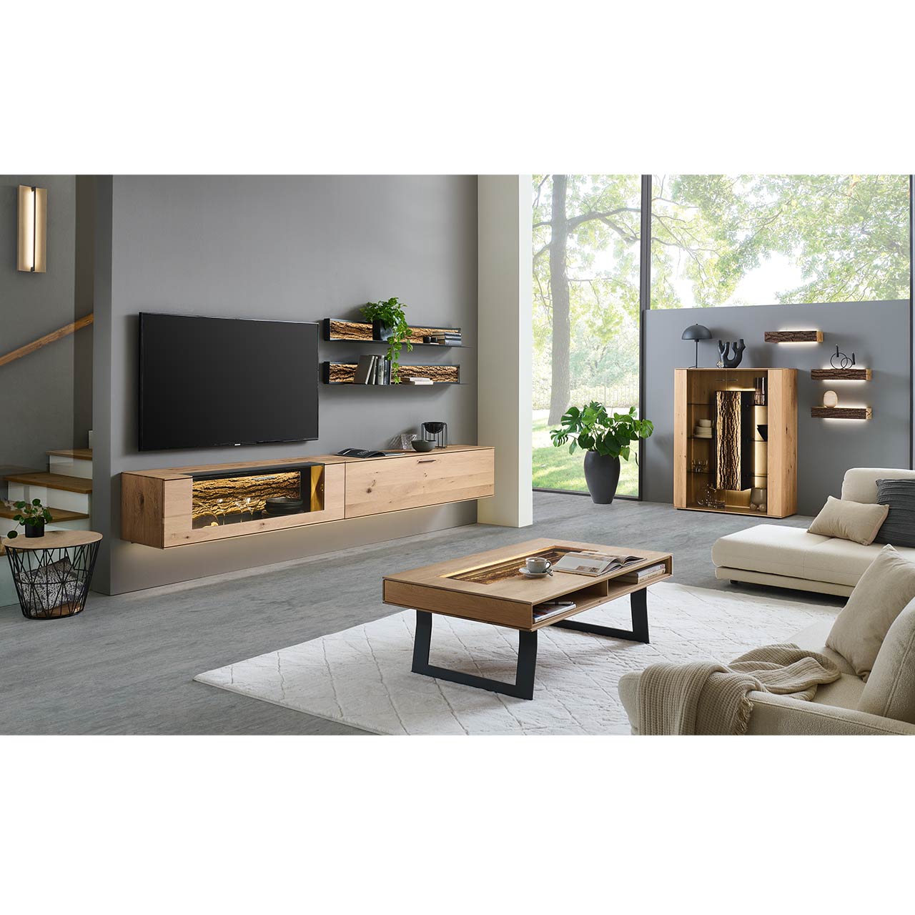 Modernes Wohnzimmer mit Wohnwand bestehend aus zwei Hängelowboards aus massiver Kerneiche Natur, eines mit beleuchtetem Glasausschnitt und Rinden-Akzent, dazu zwei Metall-Wandregale. Rechts passendes Highboard, davor Holzcoutisch.