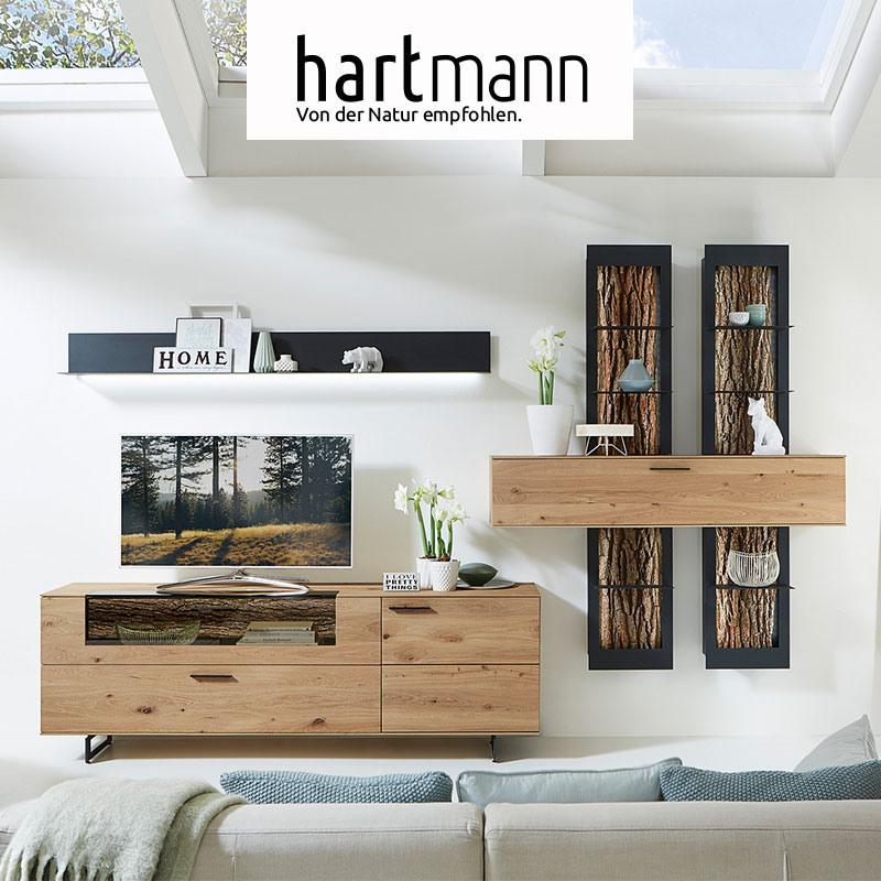Hartmann RUNA - Massivholzmöbel aus Kerneiche Natur kombiniert mit echter Baumrinde und Metall anthrazit. Ambientebild zeigt Lowboard mit Glasklappe, darüber hängend ein Metallregal, rechts ein Holzhängeschrank an zwei Wandpaneelen mit edler Baumrinde.
