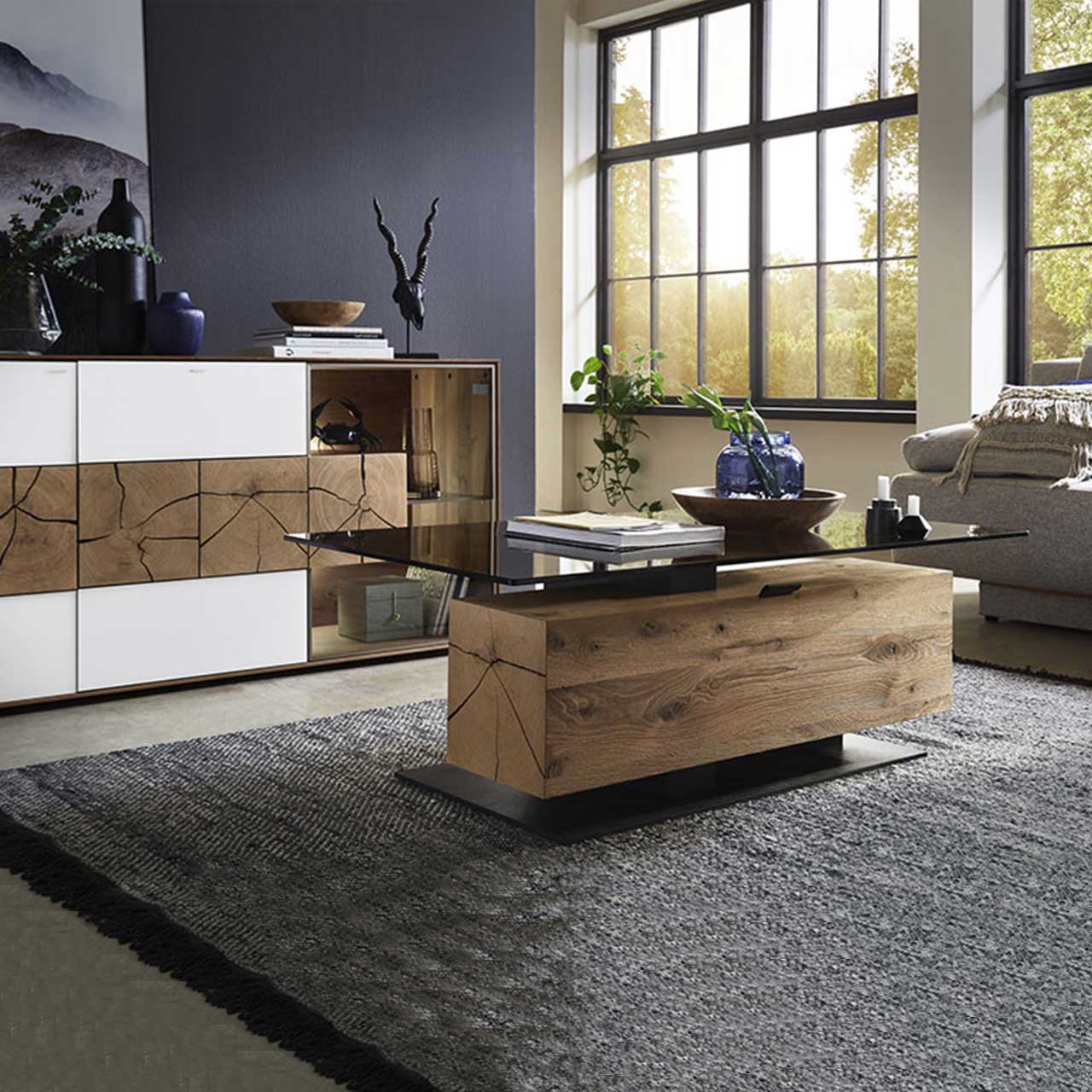 Bildausschnitt eines Wohnzimmers mit modernem Lowboard aus massiver Kerneiche Umato mit weißfarbenen Mattglasfronten, drei rechteckigen Hirnholzscheiben und Glastür. Davor passender Couchtisch mit Glasplatte und Holzblock mit Stauraumfach.
