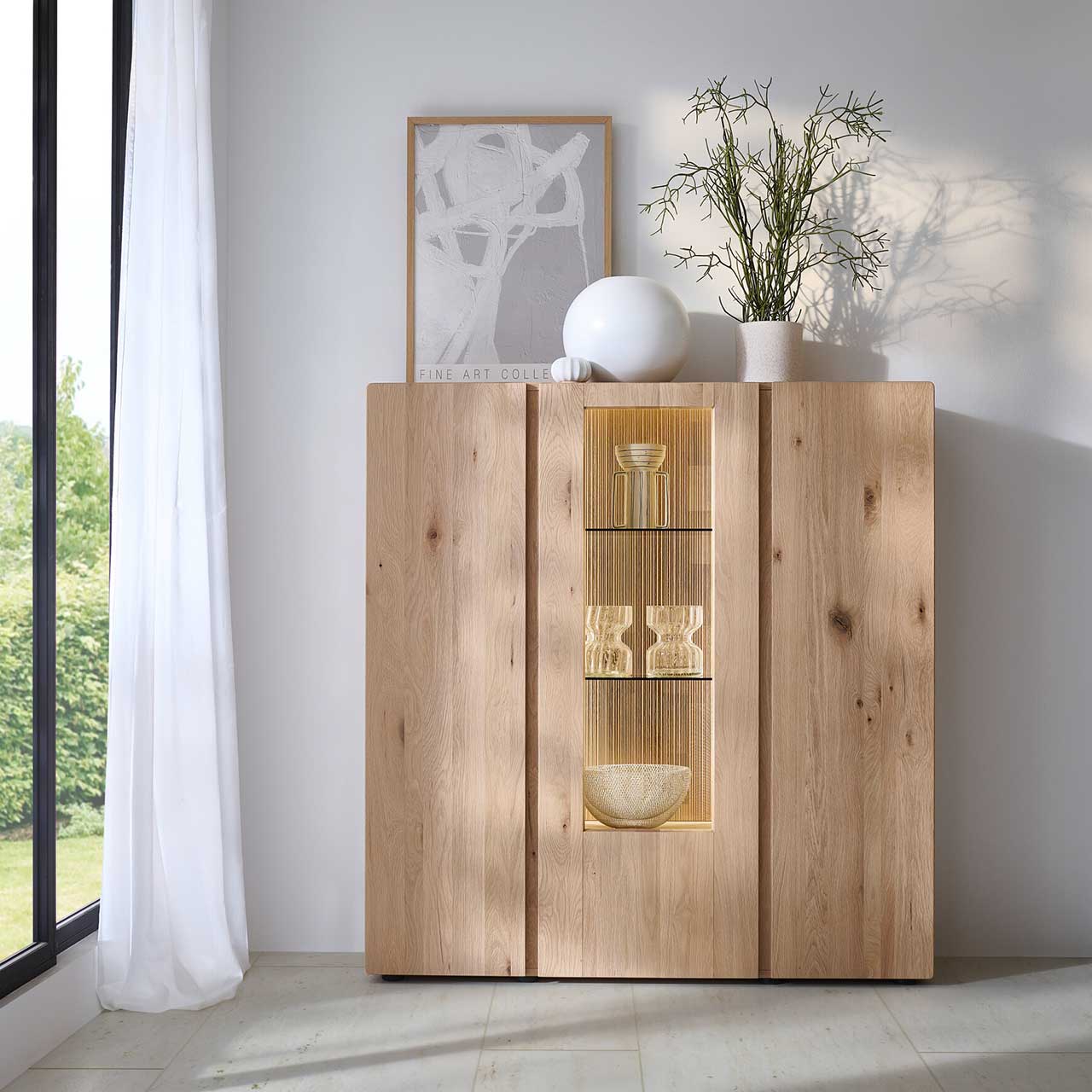 Milieubild - Hartmann VIDA - Highboard 8630-7141 - Kerneiche Natur / Glas Modernes 3-türiges Highboard aus massiver Kerneiche Natur mit zwei Holztüren rechts und links und mittig mit einer beleuchteten Glas-/Holztür, dahinter sichtbar die edle geschoppte Rückwand - in Frontansicht an einer hellgrauen Wand stehend.