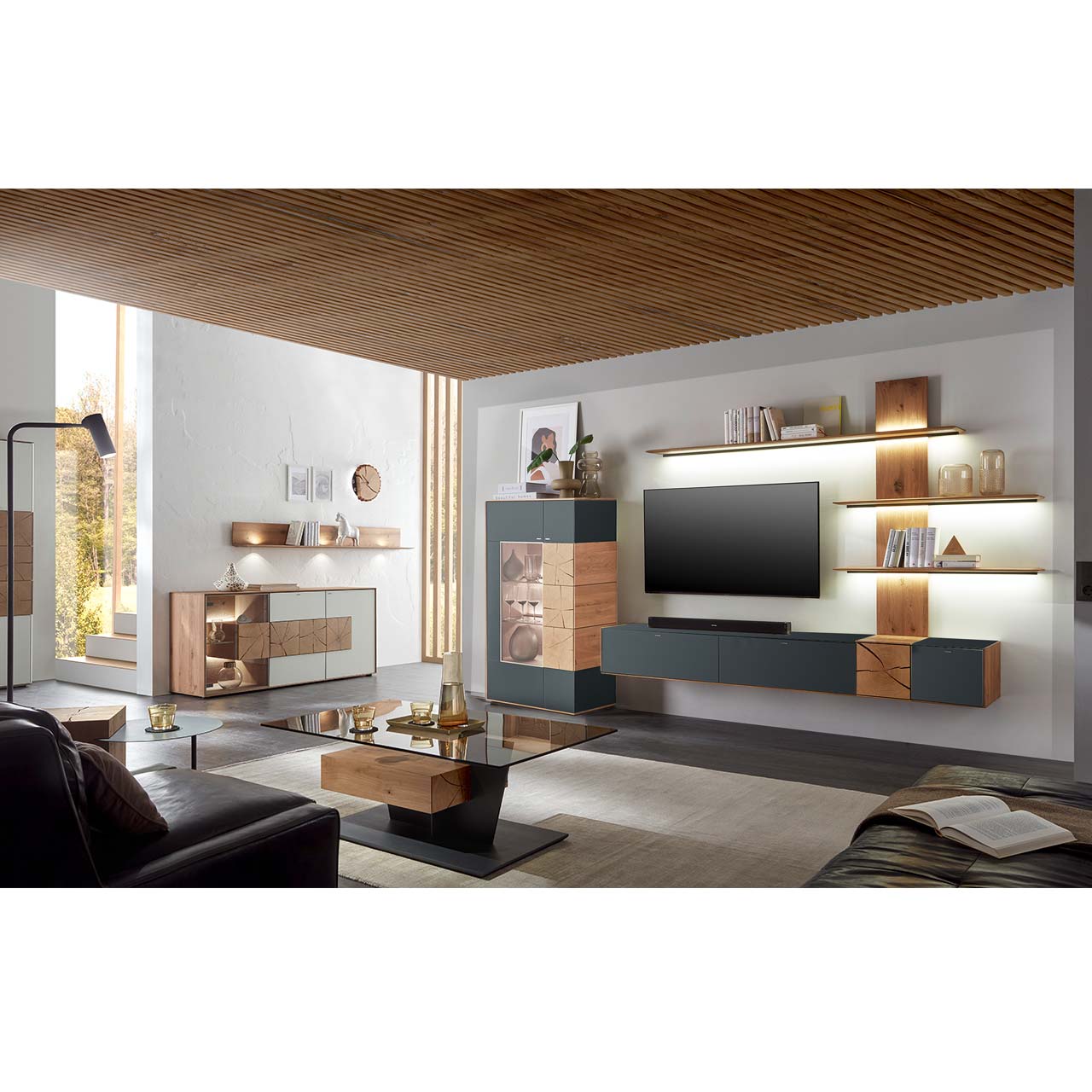 Wohnzimmer mit TV und moderner Wohnwand aus massiver Kerneiche und Mattglas anthrazit Fronten, mit schmalem Hängelowboard, beleuchtetem Wandpaneel und Standvitrine. Rechts ein Highboard darüber Regal. Im Vordergrund ein Couchtisch mit Glasplatte.