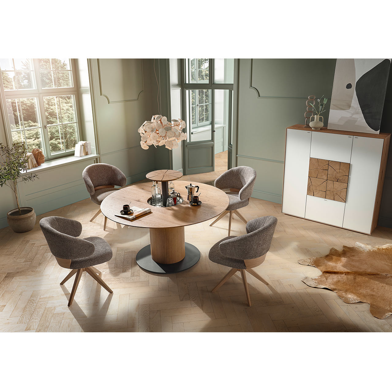 Modernes Esszimmer mit rundem Esstisch aus hellem Holz mit praktischer Flaschenaufbewahrung in der Tischmitte.