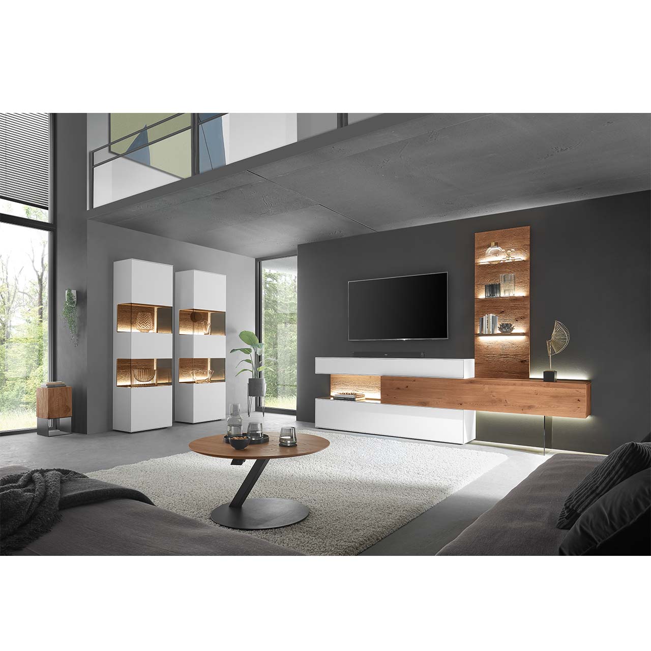 Wohnzimmer mit moderner Wohnwand aus weiß lackierter Birke und dunkler Kerneiche, bestehend aus Hängelowboard, Ober- und Unterschrank mit Beleuchtung sowie Wandpaneel mit Glasboden. Links zwei weiße Standvitrinen, davor ein runder Couchtisch.