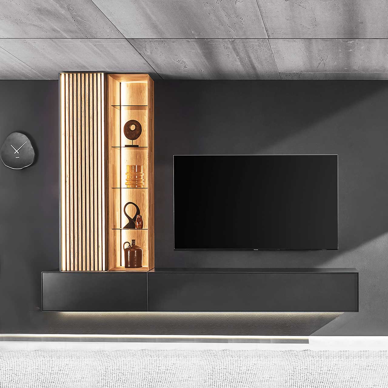 Wohnzimmer mit Fernseher und moderner, vierteiliger Wohnwand aus massivem Holz: zwei anthrazit lackierte Hängelowboards, links darauf ein Hochschrank mit Lamellenfront und ein Wandregal mit Glasböden – vor dunkelgrauer Wand