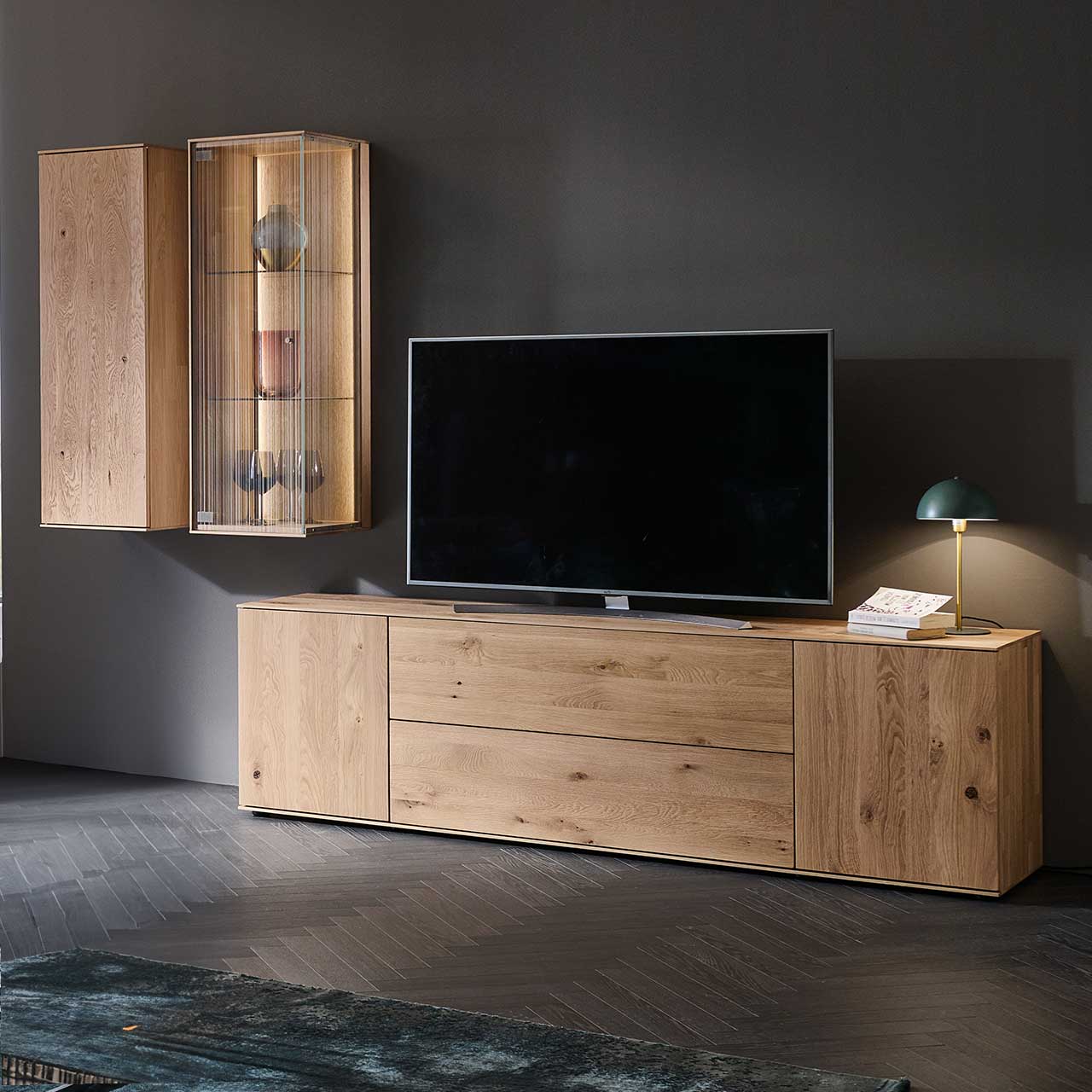 LINEA Leonardo Living - Wohnwand 8540 Nr. SV22 Wohnzimmer mit Fernseher und moderner dreiteiliger Wohnwand aus Kerneiche Natur: Eine Hängevitrine mit satiniertem Glas, ein Hängeschrank und ein schlichtes Lowboard mit Holztüren und Schubkasten – platziert vor einer dunkelgrauen Wand.