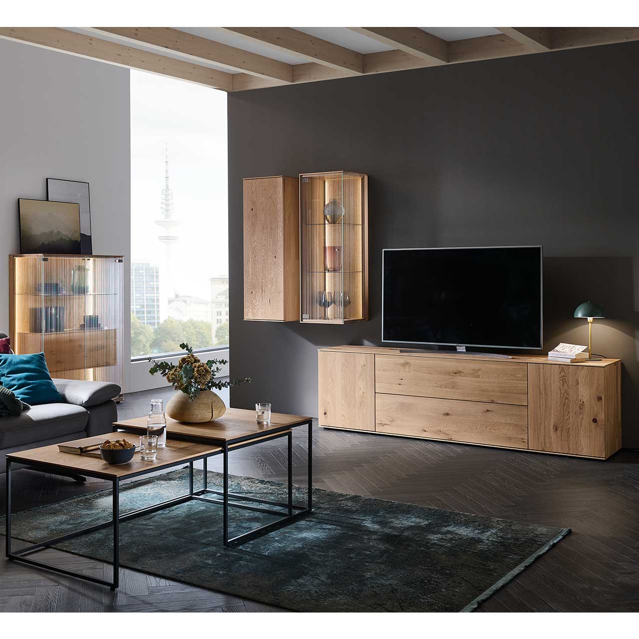 Modernes Wohnzimmer mit dreiteiliger Wohnwand aus Kerneiche Natur: Hängeschrank, Hängevitrine und Lowboard vor dunkelgrauer Wand. Links an der Wand ein Highboard mit Glastüren, im Vordergrund zwei ineinander schiebbare Couchtische.
