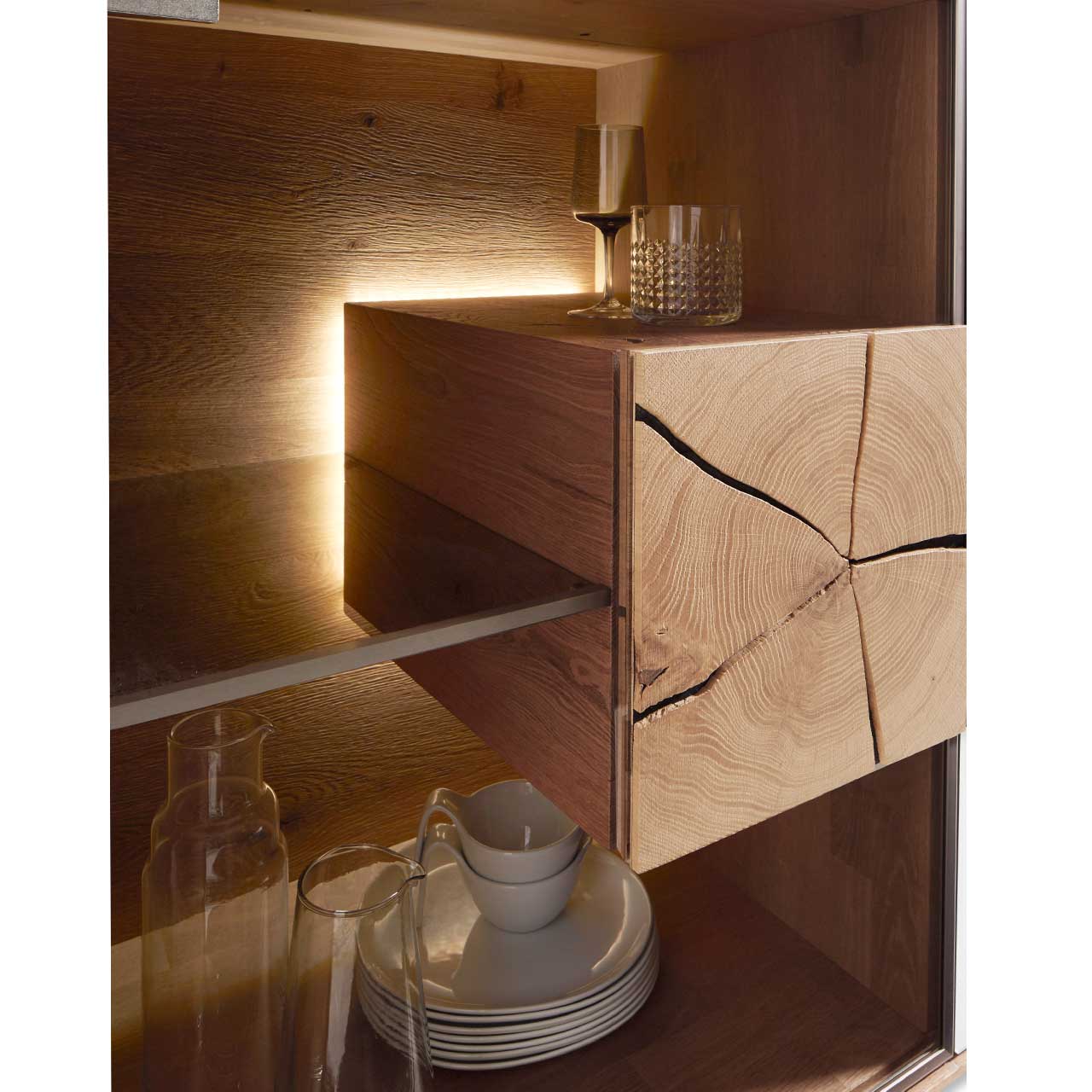 Hartmann CAYA - 7170 Detailbild Vitrinenbeleuchtung Detailbild eines beleuchteten Hirnholzblocks aus massiver Kerneiche Umato mit sichtbarer Holzmaserung und charakteristischen Holzrissen an Glastür eines CAYA Lowboards.