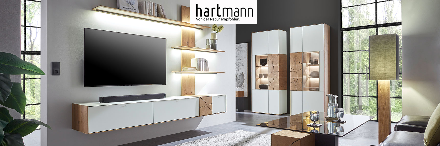 Hartmann CAYA - Massivholzmöbel aus Kerneiche Umato mit Mattglas weiß oder anthrazit Fronten und dekorativen Hirnholzscheiben. Ambientebild zeigt Hängelowboard weiß, beleuchtetes Wandpaneel aus Holz und zwei Standvitrinen mit Hirnholzscheiben.