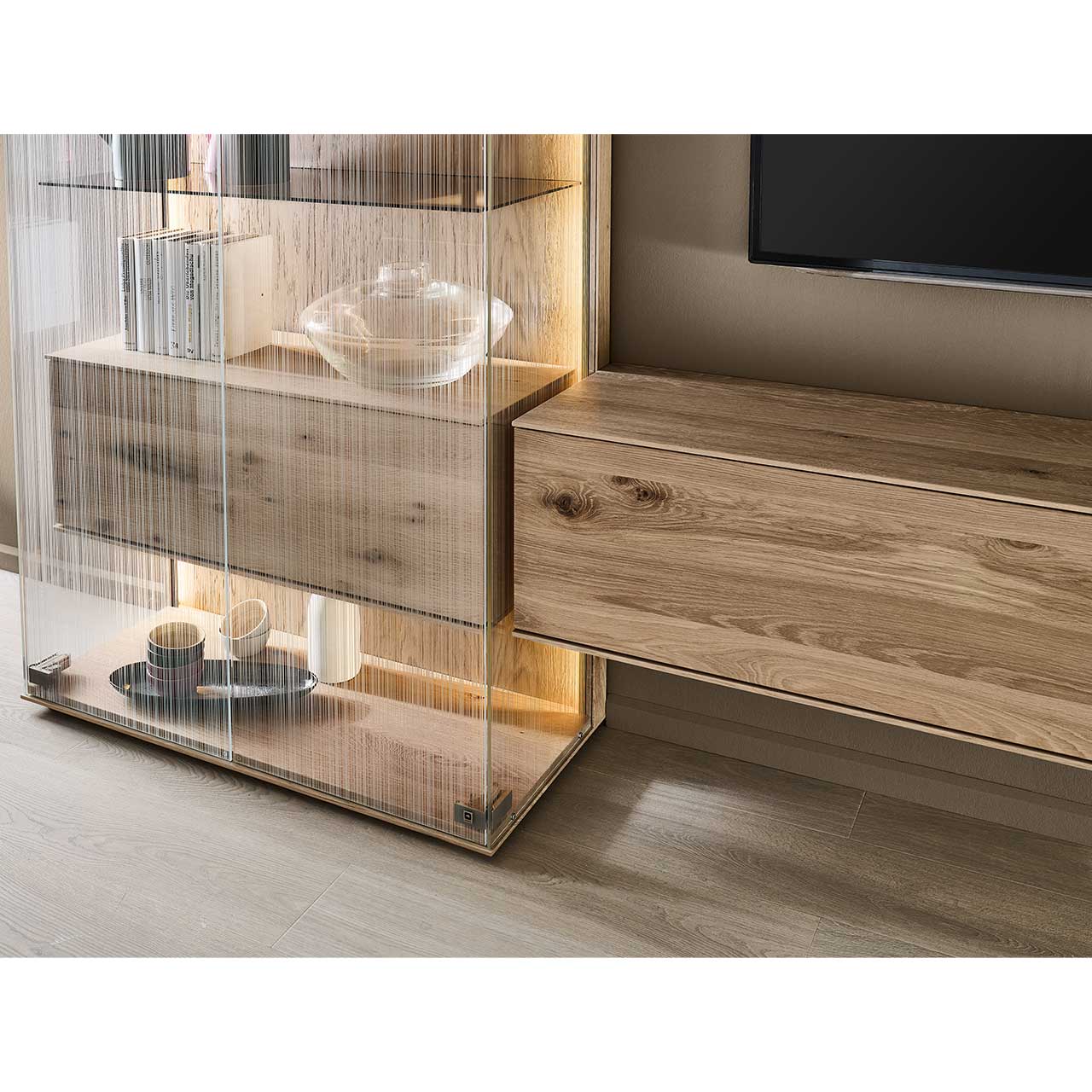 Detailaufnahme eines beleuchteten Highboards mit satinierten Glastüren und innenliegendem Holzkorpus mit Klappe. Daneben schlichtes Hängelowboard – beide aus Kerneiche Natur.