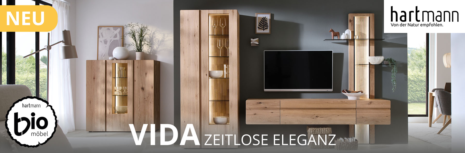 Hartmann Möbel - Möbelserie VIDA - Wohn-/Esszimmermöbel aus Kerneiche Natur Hartmann VIDA - Massivholzmöbel aus Kerneiche Natur kombiniert mit geschroppten Rückwänden und Holzeinkerbungen. Ambientebild zeigt Hängelowboard integriert in ein Wandpaneel mit Glasböden, links eine Standvitrine. Hinten links passendes Highboard.